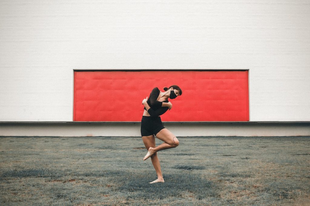 Personal development through the pursuit of passion and purpose, Full body of flexible barefoot female dancer wearing protective mask standing on one leg while performing dance movement on street